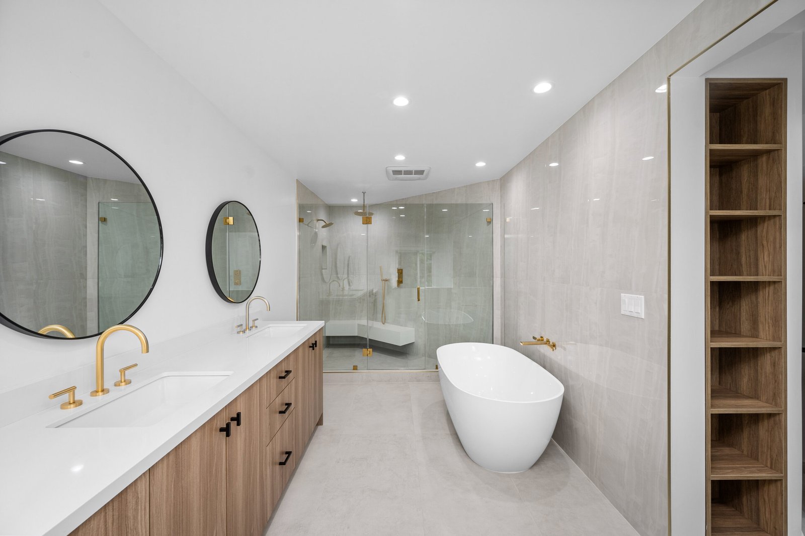 Modern bathroom with double vanity, round mirrors, brass fixtures, freestanding tub, glass walk-in shower, and built-in wood shelving.